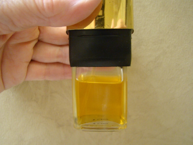 Puede incluir: Una peque&ntilde;a botella de vidrio transparente llena de un l&iacute;quido amarillo dorado. La botella tiene una banda negra y una tapa dorada. La botella est&aacute; sujeta por una mano.