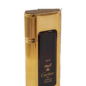May include: A gold bottle of Must de Cartier perfume. The bottle is rectangular with a gold cap and a black label that reads 