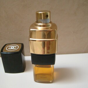Puede incluir: Un frasco de perfume dorado con una banda negra y un tap&oacute;n negro a juego. El frasco tiene forma rectangular con un tap&oacute;n dorado y un cuerpo de vidrio transparente que contiene un l&iacute;quido &aacute;mbar claro. El tap&oacute;n presenta un logotipo dorado.