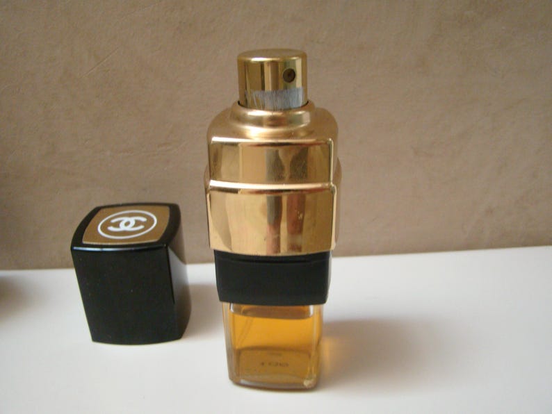 Puede incluir: Un frasco de perfume dorado con una banda negra y un tap&oacute;n negro a juego. El frasco tiene forma rectangular con un tap&oacute;n dorado y un cuerpo de vidrio transparente que contiene un l&iacute;quido &aacute;mbar claro. El tap&oacute;n presenta un logotipo dorado.