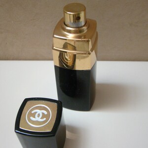 Puede incluir: Un frasco de perfume negro y dorado con una tapa a juego. El frasco tiene forma rectangular con una parte superior dorada y una base negra. La tapa presenta un borde dorado y un logotipo blanco.