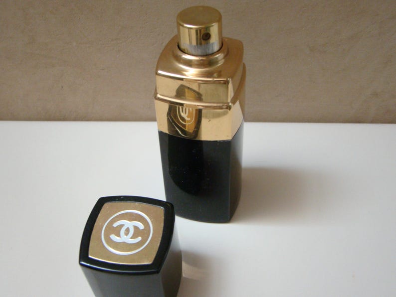 Puede incluir: Un frasco de perfume negro y dorado con una tapa a juego. El frasco tiene forma rectangular con una parte superior dorada y una base negra. La tapa presenta un borde dorado y un logotipo blanco.