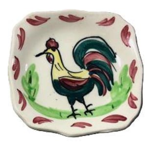 May include: A small, square ceramic dish with a hand-painted rooster design. The rooster is depicted in black, yellow, green, and red. The dish has a white background with red leaf-like accents around the rim.