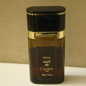 May include: A small bottle of Must de Cartier perfume. The bottle is black with gold accents and a clear glass body. The label reads 