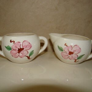 May include: A cream-colored ceramic sugar bowl with two handles and a matching creamer pitcher, both featuring a hand-painted pink flower design with green leaves and brown stems. The set has a smooth, glossy finish and rounded shapes.