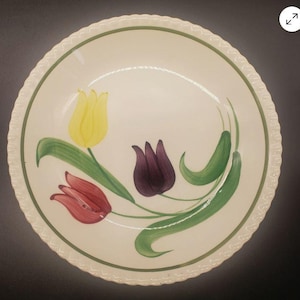 May include: A round, cream-colored ceramic bowl featuring a hand-painted design of three tulips in yellow, red, and purple with green stems and leaves. The bowl has a scalloped edge and a thin green circular border painted along the inner rim.