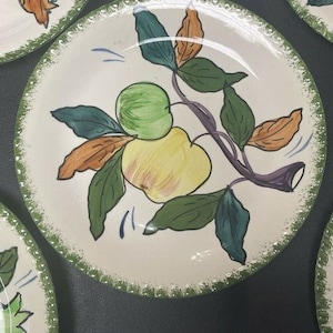 May include: A round ceramic plate with an off-white base and a green speckled rim. The center features a hand-painted illustration of a green apple and a yellow apple on a branch with dark green and brown leaves, accented by thin blue lines.