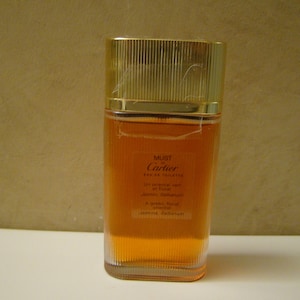 May include: A bottle of Must de Cartier Eau de Toilette perfume. The bottle is clear glass with a gold cap and a gold label. The label has the words 