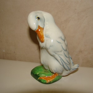 May include: A ceramic duck figurine with a glossy off-white finish, featuring a bright orange beak and feet. The duck is depicted in a preening pose, standing on a small, textured green base with subtle blue-grey feather detailing on its wings.