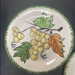 May include: A round ceramic plate with a cream-colored base and a hand-painted design of yellow grapes, green leaves, and a single brown leaf. The rim features a textured green border with a dotted pattern, framing the central fruit illustration.