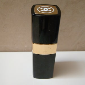Puede incluir: Barra de labios negra con una banda dorada y una parte superior dorada con un logotipo blanco. La barra de labios est&aacute; de pie sobre una superficie blanca. El fondo es de color beige neutro.