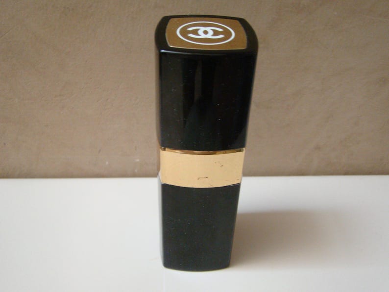 Puede incluir: Barra de labios negra con una banda dorada y una parte superior dorada con un logotipo blanco. La barra de labios est&aacute; de pie sobre una superficie blanca. El fondo es de color beige neutro.