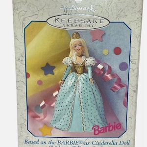 May include: A Hallmark Keepsake Ornament featuring a Barbie doll dressed as Cinderella. The doll wears a light blue gown with gold accents and a crown. The box has text that reads "Barbie" and "Collector's Series."