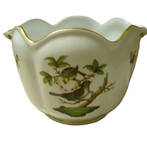 May include: A white porcelain bowl with a scalloped rim and gold trim. The exterior features a hand-painted illustration of two birds perched on a brown branch with green leaves, set against a small patch of green grass, with small butterflies.
