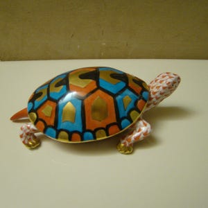 May include: A ceramic turtle figurine with a colorful shell. The shell features a geometric pattern in blue, orange, gold, and black. The turtle's head and legs are white with red and orange spots, and the feet are gold.