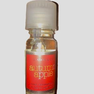 May include: A small clear glass bottle of Slatkin & Co. Autumn Apple home fragrance oil with a white screw-top cap. The red rectangular label features the text Slatkin & Co. autumn apple HOME FRAGRANCE 0.33 Fl. Oz./9.7 ml in yellow and white.