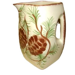 May include: Cream-colored ceramic pitcher with a brown handle and rim. The pitcher is decorated with brown pine cones and green pine needles. The pitcher has a unique, curved shape and a spout for pouring.