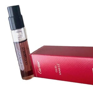 May include: A clear glass perfume vial with a black cap, containing a brown liquid, next to a red box. The vial and box both have the Cartier logo and the words "OUD VANILLE" printed on them.