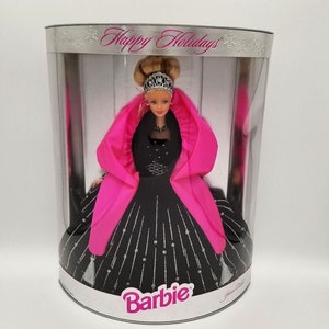 May include: A Barbie doll in a cylindrical display box labeled Happy Holidays. The doll wears a black gown with silver glitter accents, a bright pink shawl, and a silver tiara. The doll has blonde hair styled in an updo and is positioned inside the clear plastic packaging with silver metallic top and bottom rims.