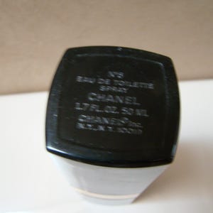 Puede incluir: Botella de perfume en blanco y negro con el texto "N&deg;5 Eau de Toilette Spray CHANEL 1.7 FL.OZ. 50 ML". La botella tiene forma rectangular con base blanca y parte superior negra.
