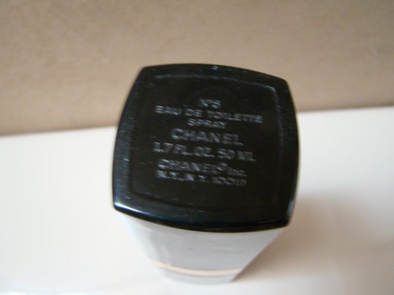 Puede incluir: Botella de perfume en blanco y negro con el texto "N&deg;5 Eau de Toilette Spray CHANEL 1.7 FL.OZ. 50 ML". La botella tiene forma rectangular con base blanca y parte superior negra.