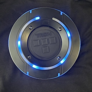 May include: Black hoodie with a circular, glowing blue light-up disc attached to the back. The disc has the word "TRON" printed on it and a series of square shapes. The hoodie is made of a soft material.