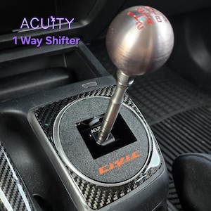 May include: A silver spherical gear shift knob with red gear pattern markings atop a metal shaft. The base features a black carbon fiber-style trim with the orange text CIVIC and a small black component labeled ACUITY. Text reads ACUITY 1 Way Shifter.