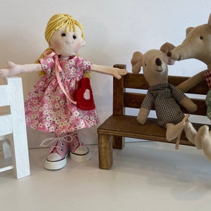 May include: A handmade doll with blonde yarn hair and a pink floral dress stands near a miniature white bench. Two stuffed animal figures, a koala and a mouse, sit on a wooden bench. The mouse wears a green floral dress.