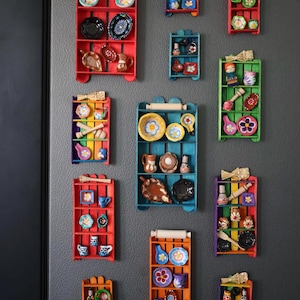 Può includere: Diverse piccole mensole in legno colorate, ognuna con piccoli oggetti in ceramica. Le mensole sono dipinte in rosso, blu, verde e arancione. Contengono piatti, ciotole, tazze e oggetti decorativi in miniatura, creando un'esposizione vivace.