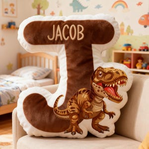 Op de afbeelding: Een pluche kussen in de vorm van de letter "J" met de naam "JACOB" in beige geborduurd. Het kussen heeft een bruin en wit kleurenschema en een illustratie van een Tyrannosaurus Rex.