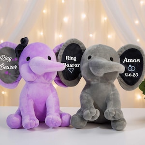 Könnte beinhalten: Zwei Ringträger-Spielzeuge in Form von Plüschelefanten. Einer ist lila, der andere grau. Jeder hat große Ohren mit dem Text "Ring Bearer" und "Amos 4-6-25". Der lila Elefant hat eine schwarze Schleife.