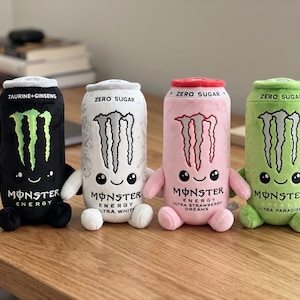 May include: Four plush toys shaped like energy drink cans in black, white, pink, and green. Each features the Monster Energy logo, a smiling face, small arms, and legs. The cans are labeled with text including Taurine+Ginseng, Zero Sugar, Ultra White, Ultra Strawberry Dreams, and Ultra Paradise.