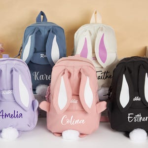Peut inclure: Cinq sacs à dos colorés sur le thème du lapin. Chaque sac à dos a de grandes oreilles de lapin et une queue touffue. Les sacs à dos sont dans des tons de bleu, lavande, rose, crème et noir. Chaque sac à dos porte un nom.