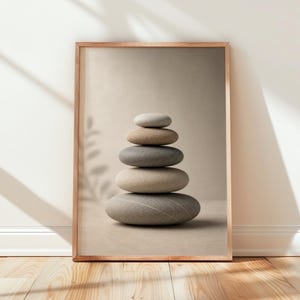 May include: A rectangular framed wall art print featuring a vertical stack of five smooth, rounded river stones in varying shades of beige, tan, and gray. The stones are balanced on top of each other, centered within the frame against a neutral background.