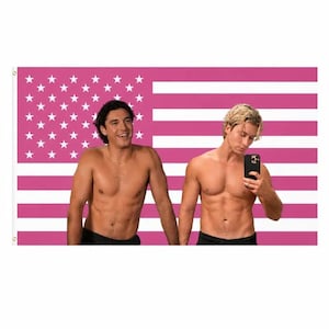 May include: A pink American flag with white stars and stripes is the backdrop for two shirtless individuals. One is smiling, and the other is holding a black phone, possibly taking a selfie. Both are wearing black shorts.