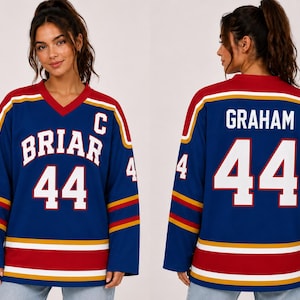 Può includere: Una maglia da hockey blu con il numero 44 in bianco. La maglia presenta dettagli rossi e oro sulle maniche e sul colletto. Il davanti dice "BRIAR" e il retro "GRAHAM".
