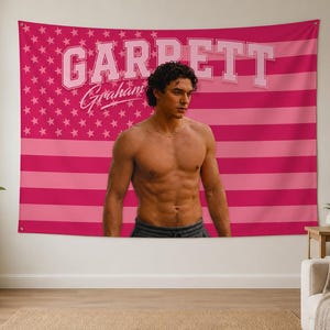 Può includere: Un arazzo rosa con un disegno della bandiera americana con il nome "Garrett Graham" in lettere bianche. Una persona senza maglietta è in piedi davanti all'arazzo. La bandiera ha strisce rosa e un campo di stelle.