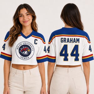 Puede incluir: Camiseta corta blanca con detalles azules y dorados. Presenta el logotipo de Briar University Hawks en la parte delantera y el nombre "GRAHAM" y el número "44" en la parte trasera. El número "4" está en cada manga.