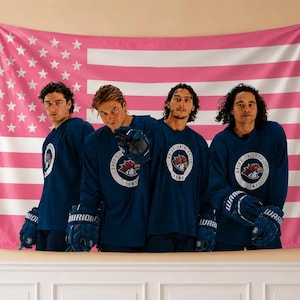 May include: A pink American flag tapestry hangs behind four people wearing navy blue hockey jerseys. The jerseys have a circular logo and the words "Brian University Hawks". They are wearing blue hockey gloves.