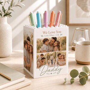 May include: A white pen holder with a photo collage and the text "We Love You Daddy". The pen holder contains five pens in pastel colors: green, blue, purple, orange, and pink.
