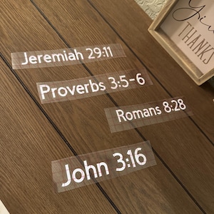 May include: A wooden surface with four transparent stickers displaying Bible verses: Jeremiah 29:11, Proverbs 3:5-6, Romans 8:28, and John 3:16. A framed sign with the words "Give Thanks" is in the background.