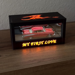 May include: A red classic car model inside a black display case with the text "MY FIRST LOVE" illuminated in yellow. The top of the case features an orange dragon design. The display case sits on a wooden surface.
