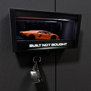 May include: A black wall-mounted display case with an orange sports car model inside. The text "BUILT NOT BOUGHT" is illuminated at the bottom. A set of keys hangs below the display case.