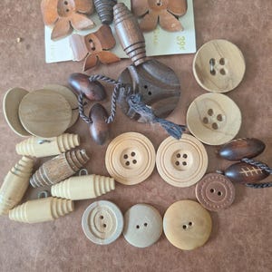 May include: Assortment of wooden buttons in various shapes and sizes, including round, oval, and bow-shaped buttons. Some buttons have a natural wood finish, while others are stained in shades of brown. The buttons are arranged on a brown surface.