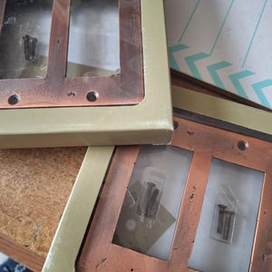 May include: Two copper-colored double light switch plates with a distressed finish, set in a gold-colored cardboard box. Each plate has two rectangular openings and includes screws. The plates are new and unused.