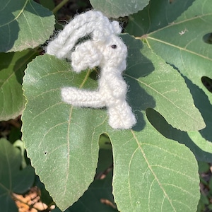 May include: A small, handmade white crochet bunny with black eyes rests on a large green leaf. The bunny is crafted with visible stitches, giving it a textured appearance. The background features more green leaves, suggesting a natural setting.
