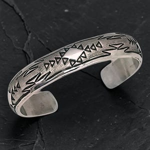 May include: A silver cuff bracelet featuring a polished finish with oxidized recessed areas. The surface is decorated with a repeating pattern of stamped triangles and a central diamond shape, creating a textured look across the metal band.