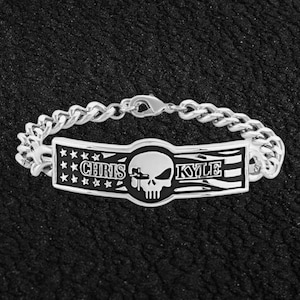 May include: Silver-toned bracelet with a curb chain design. The centerpiece features the names "CHRIS" and "KYLE" flanking a skull emblem. The design incorporates stars and stripes, with a black and silver color scheme.
