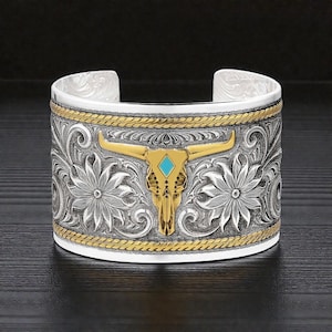 May include: Silver cuff bracelet with a gold bull skull centerpiece and turquoise stone. The bracelet features intricate floral and rope detailing. The bull skull has gold horns and a turquoise stone in the center.