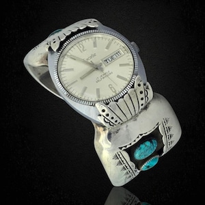 May include: A silver Excella watch with a cream face, showing the day and date. The watch is set in a decorative silver cuff with turquoise accents and engraved patterns. The watch band is approximately 5 cm wide.